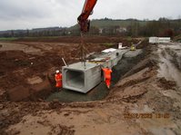 Ein Kran bringt ein Betonteil auf eine Baustelle, zwei Bauarbeiter setzen es an die richtige Stelle