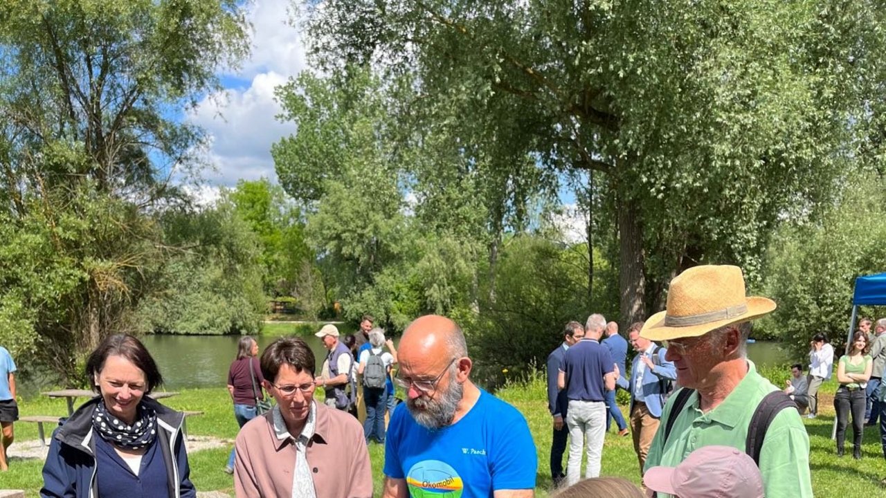 Bild zeigt Regierungspräsidentin Susanne Bay mit den Ökomobilisten an einem Tisch, im Hintergrund ein See und viele Menschen