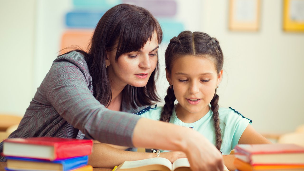 Junge Lehrerin mit Schülerin an einem Schreibtisch mit vielen Büchern darauf