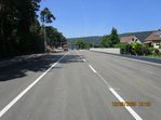 Straße, im Hintergrund drei LKW, rechts neben der Straße zwei Häuser, links stehen Bäume