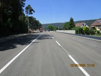 Straße, im Hintergrund drei LKW, rechts neben der Straße zwei Häuser, links stehen Bäume