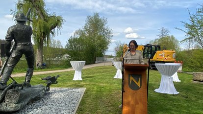 Frau Regierungspräsidentin Bay steht am Rednerpult und hält Ihre Ansprache, dahinter Stehtische und ein Bagger, neben Ihr eine Skulptur