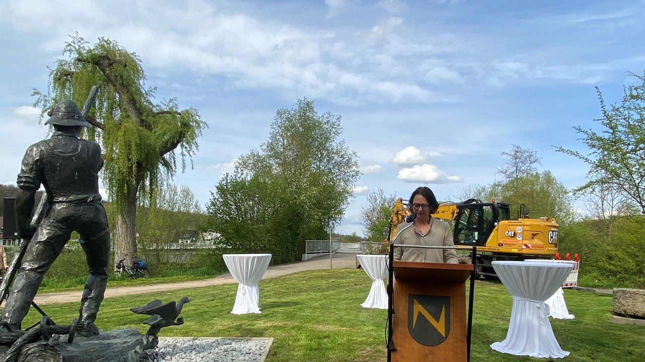 Frau Regierungspräsidentin Bay steht am Rednerpult und hält Ihre Ansprache, dahinter Stehtische und ein Bagger, neben Ihr eine Skulptur