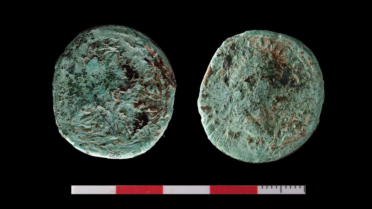 Eine Bronzemünze (unrestauriert, 1 Dupondius = ½ Sesterz) aus dem Bereich der römischen Straße. Schemenhaft ist auf der Vorderseite (rechts) das Abbild eines Kaisers (vermutlich Kaiser Domitian) zu erkennen.