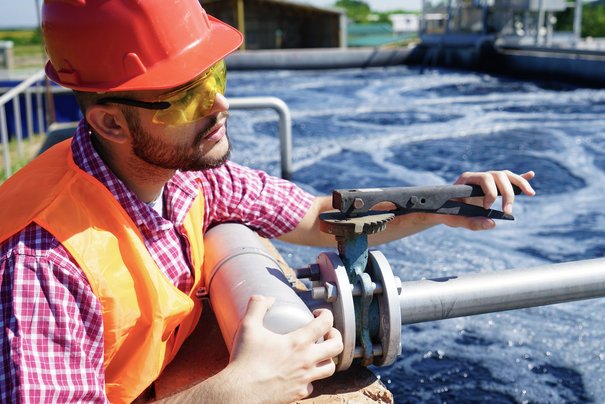 Abwassermeister arbeitet an einem Rohr, im Hinergrund sieht man die Kläranlage