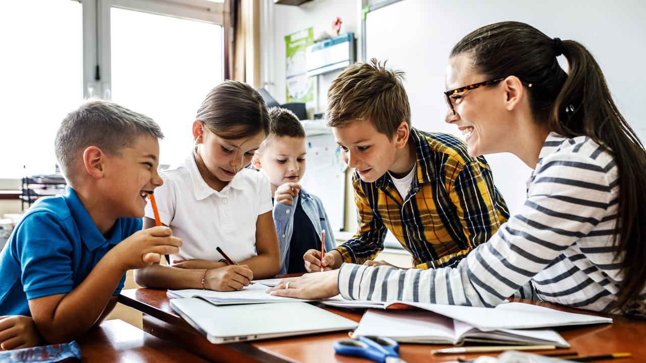 Eine Lehrerin lernt mit 4 Kindern an einem Tisch, Bücher, Hefte und Stifte liegen darauf