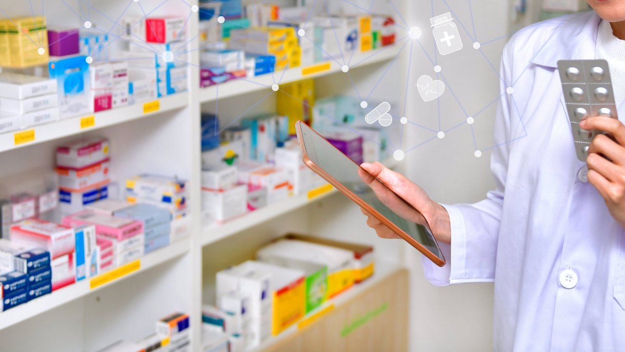 Regale mit Arzneimitteln, davor Apothekerin mit einem Tablet in der einen und Tabletten in der anderen Hand