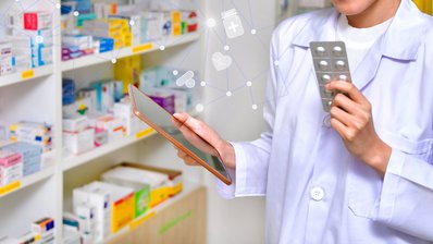 Regale mit Arzneimitteln, davor Apothekerin mit einem Tablet in der einen und Tabletten in der anderen Hand