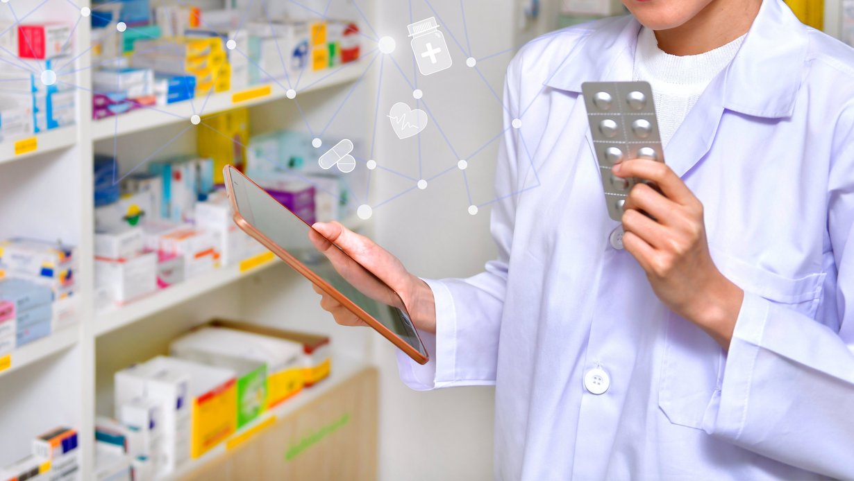 Regale mit Arzneimitteln, davor Apothekerin mit einem Tablet in der einen und Tabletten in der anderen Hand