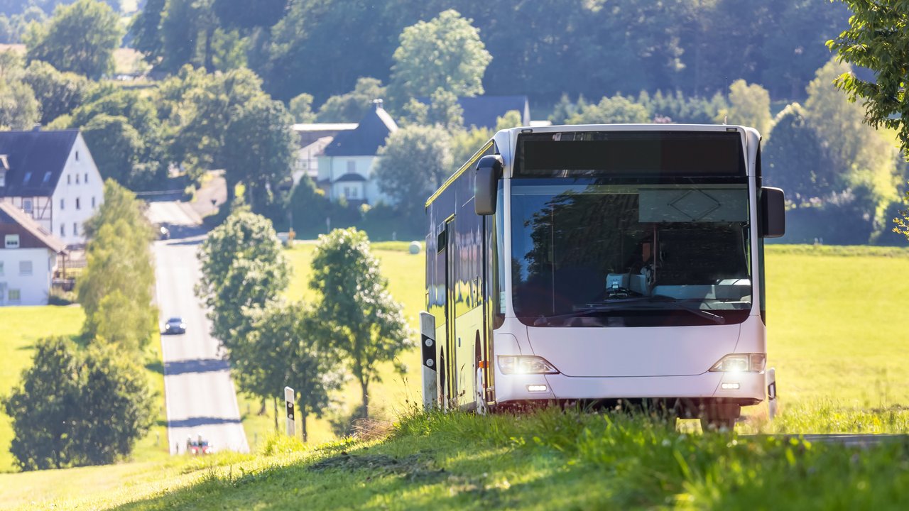 Bus fährt auf Landstraße, hinter sich eine Ortschaft und Autos