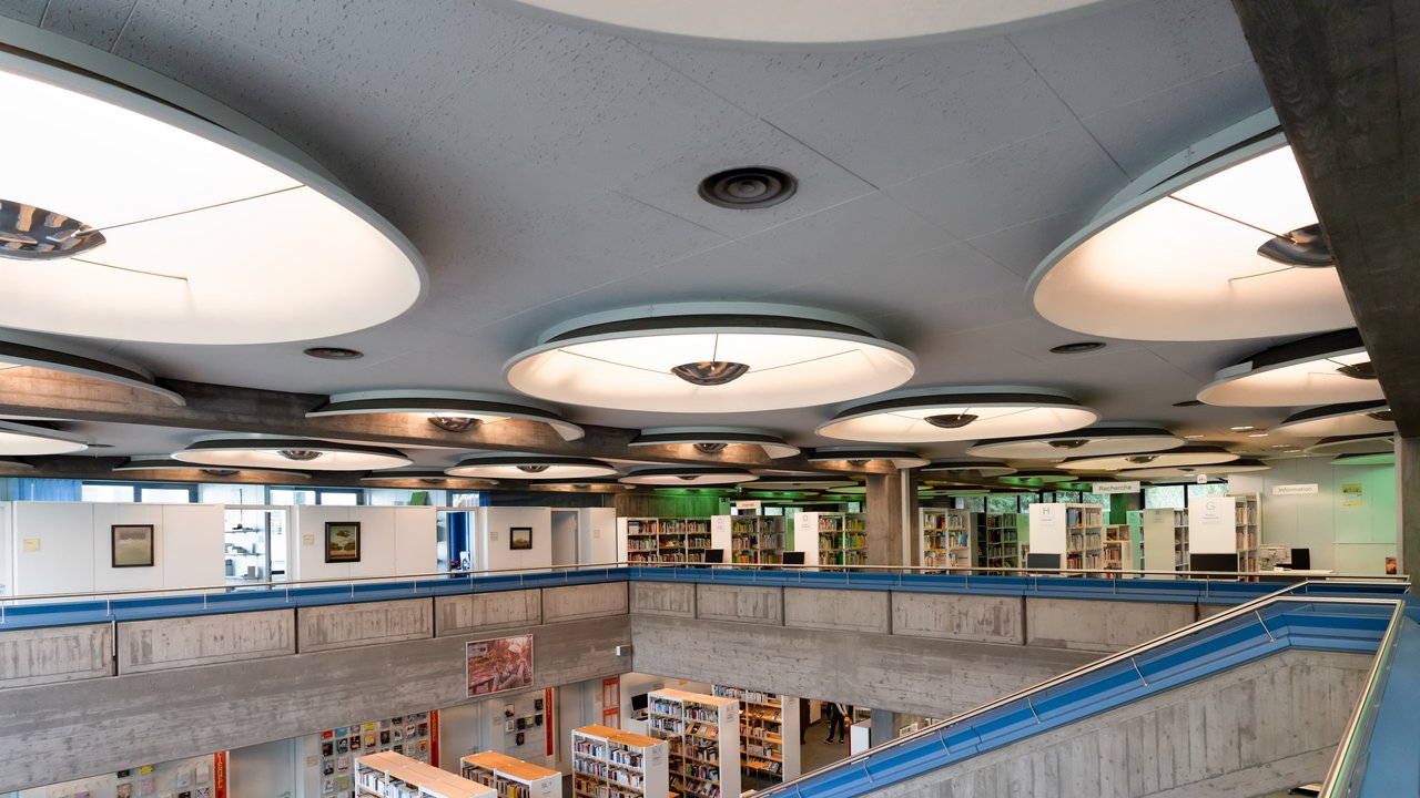 Stadtbibliothek fotografiert vom oberen Stockwerk, rechts eine Treppe, die nach unten führt, überall Bücherregale mit Büchern