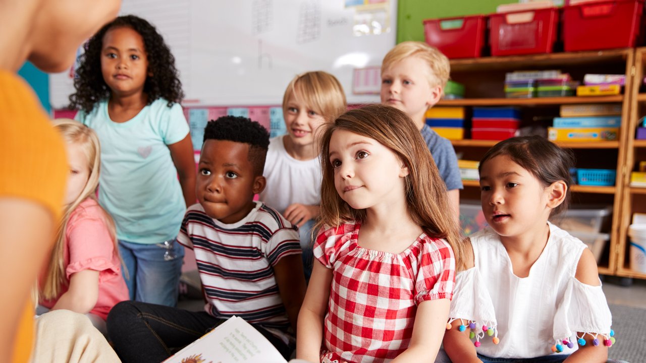 Sechs Kinder sitzen auf dem Boden vor einer Lehrerin, die vorliest