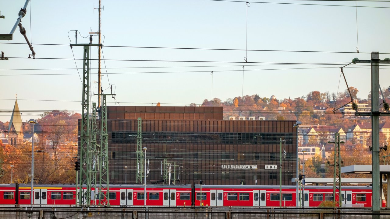 Bild zeigt eine rote S-Bahn mit Gleisen und Oberleitungen im Vordergrund und einem großen, rotem Gebäude im Hintergrund