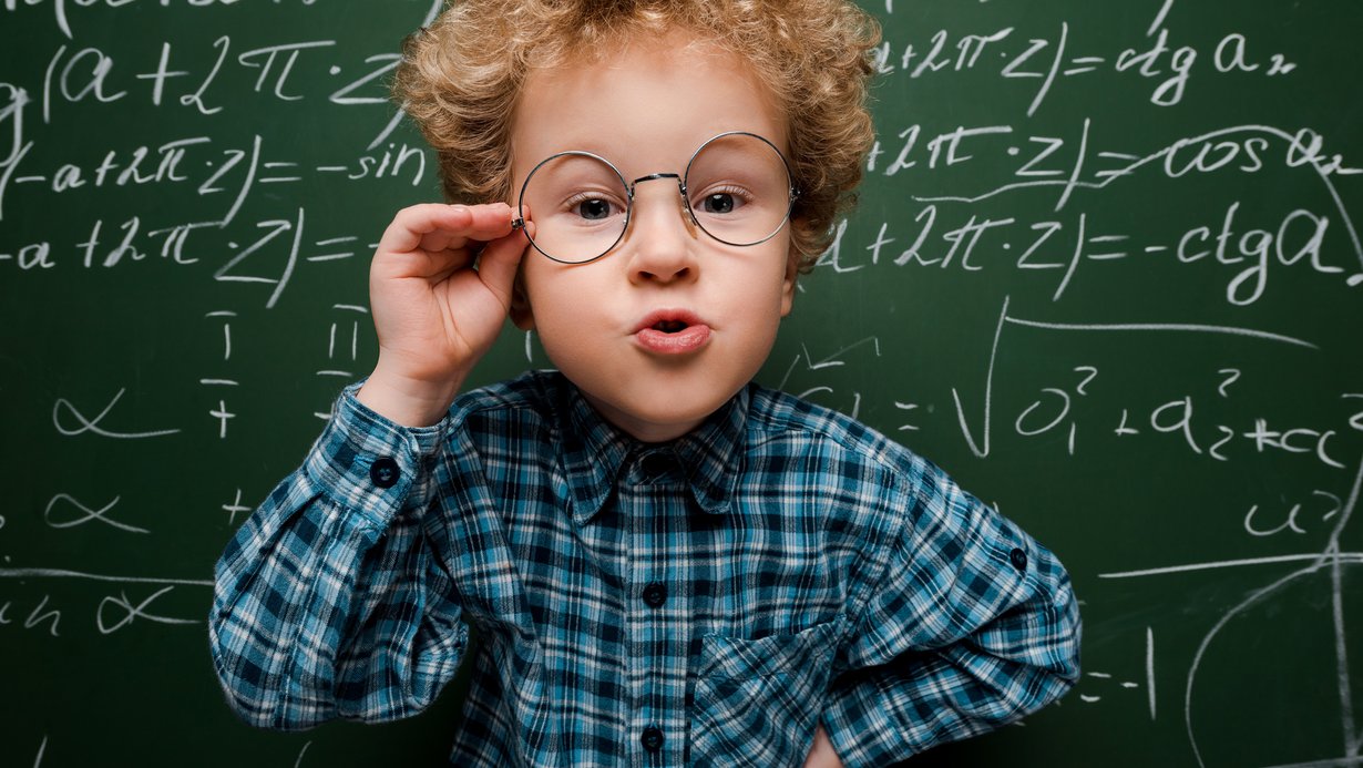 Kind, das seine Brille berührt und mit der Hand auf der Hüfte vor der Tafel steht