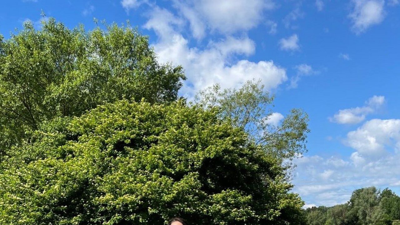 Bild zeigt Regierungspräsidentin Susanne Bay, im Hintergrund ein See, vier Kinder und eine Frau sitzen auf Bänken vor einem Tisch