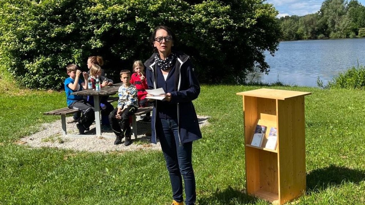 Regierungspräsidentin Susanne Bay bei der Einweihung des Bibertour-Rucksacks Bild zeigt Regierungspräsidentin Susanne Bay, im Hintergrund ein See, vier Kinder und eine Frau sitzen auf Bänken vor einem Tisch