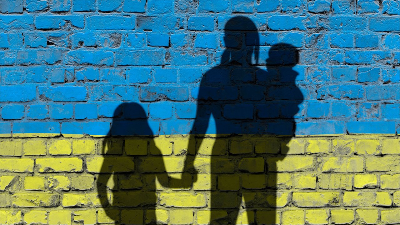 Bild zeigt Schattenumriss einer Frau mit einem Mädchen an der Hand und einem Kleinkind auf dem Arm vor einer Ziegelwand in den Farben der ukrainischen Flagge