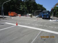 Straßenkreuszung, auf der Baufahrzeuge stehen, ein Arbeiter läuft links über die Straße, im Hintergrund Bäume