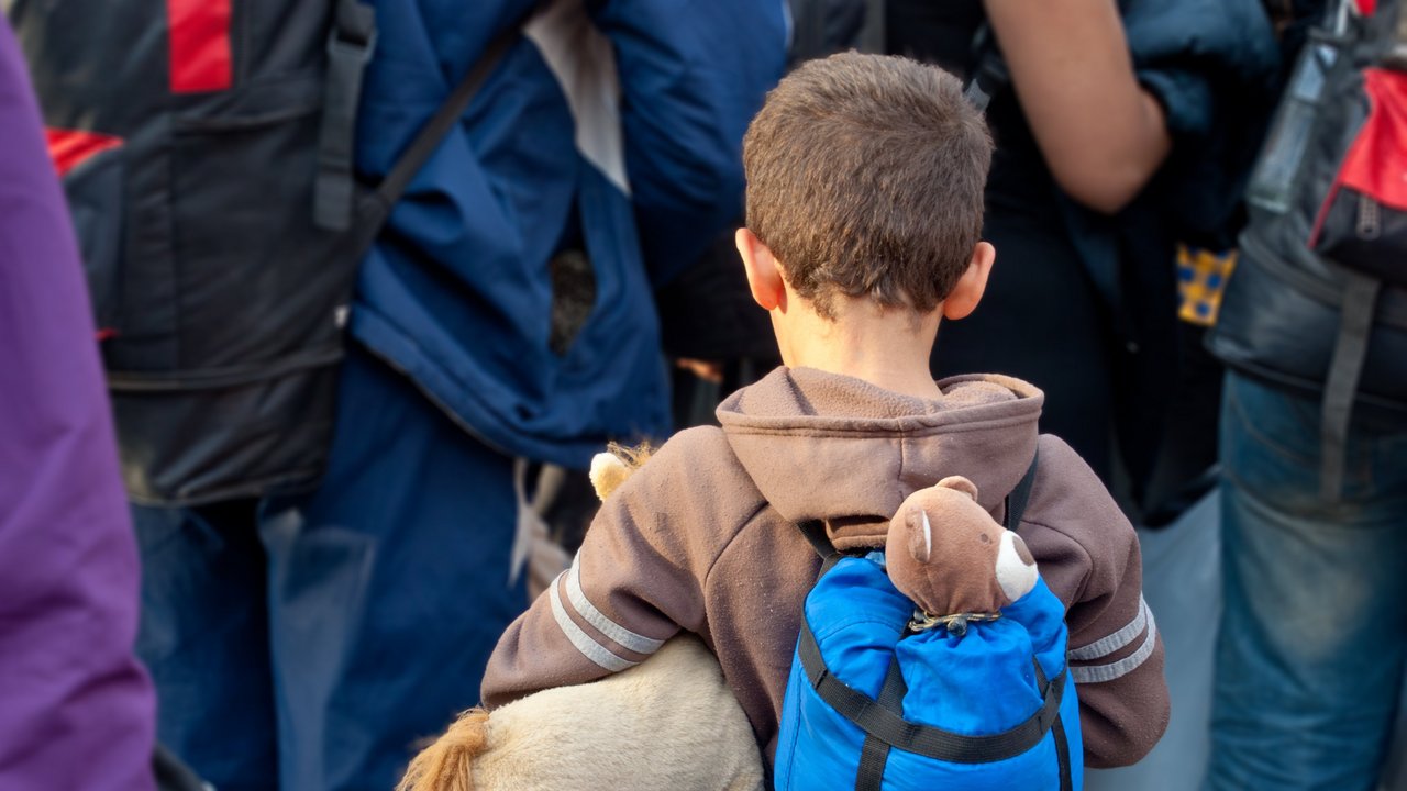 Bild zeigt Flüchtlinge, vorne ein Junge mit Rucksack, unter dem Arm ein Stofftier