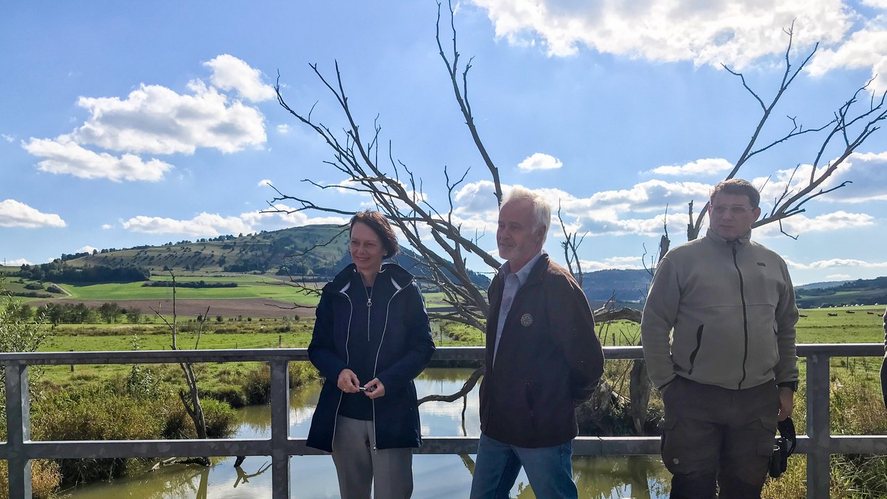 Bild zeigt Regierungspräsidentin Bay, Wolfgang Schnedier, und Armin Dammenmiller auf einer Brücke, darunter ein Bach, rechts und links Wiese und Hecken