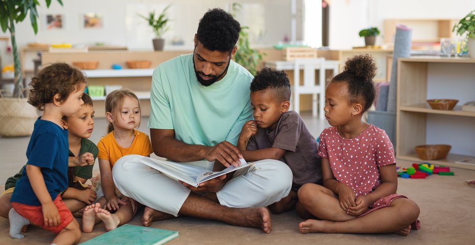 Mann liest Kindern vor, sitzend auf dem Boden, die Kinder im Halbkreis um ihn herum