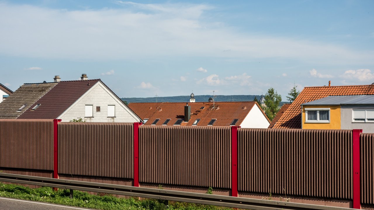 Braune Lärmschutzwand entlang einer Straße, dahinter sieht man die Gebel von Häusern