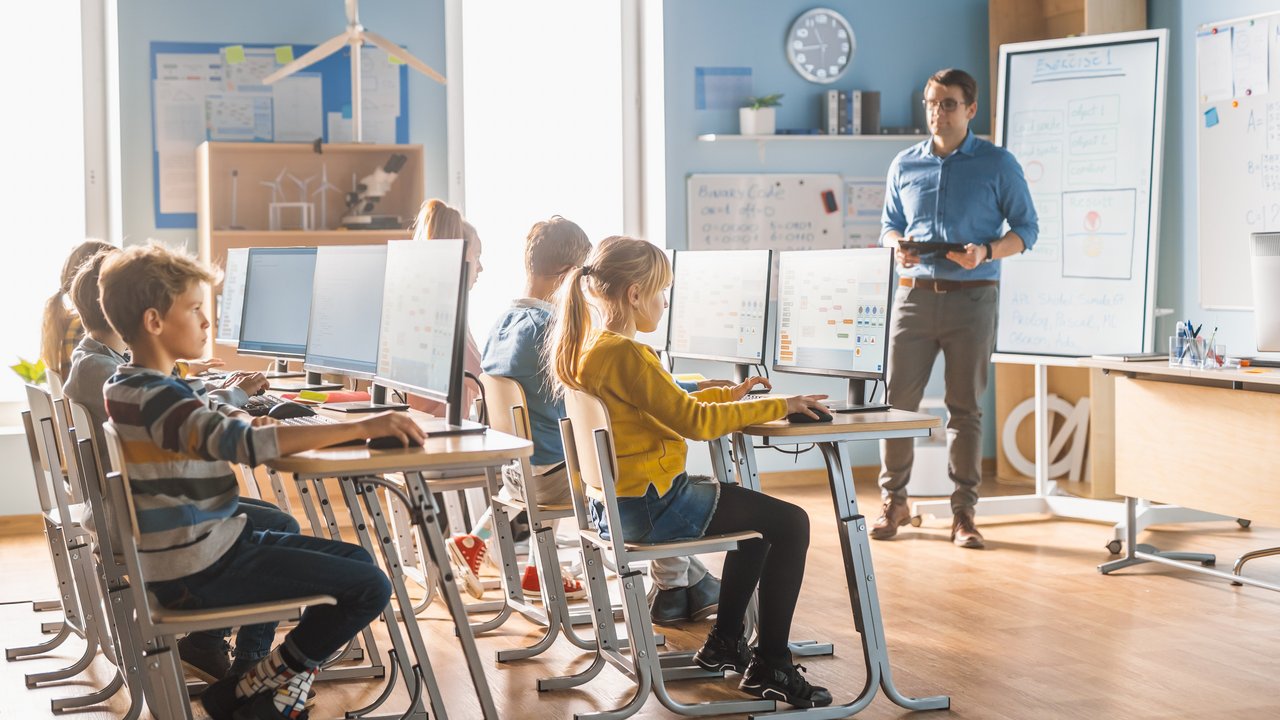 Kinder sitzen in der Schule am Computer der Lehrer steht mit dem Tablet vor ihnen