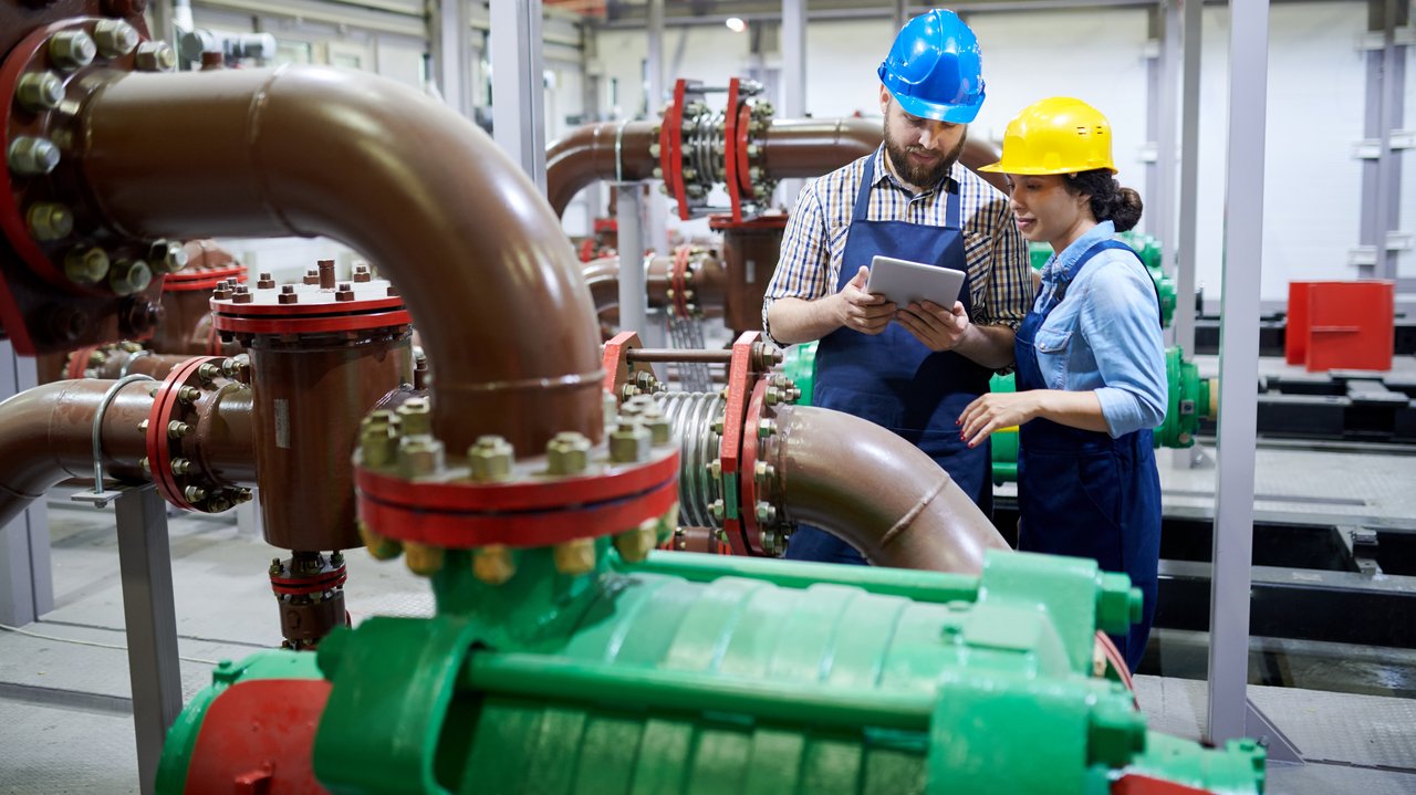Zwei Fachkräfte für Wasserversorgungstechnik, die ein digitales Tablet verwenden, während sie an Rohrleitungen und Maschinen in der Werkstatt arbeiten.