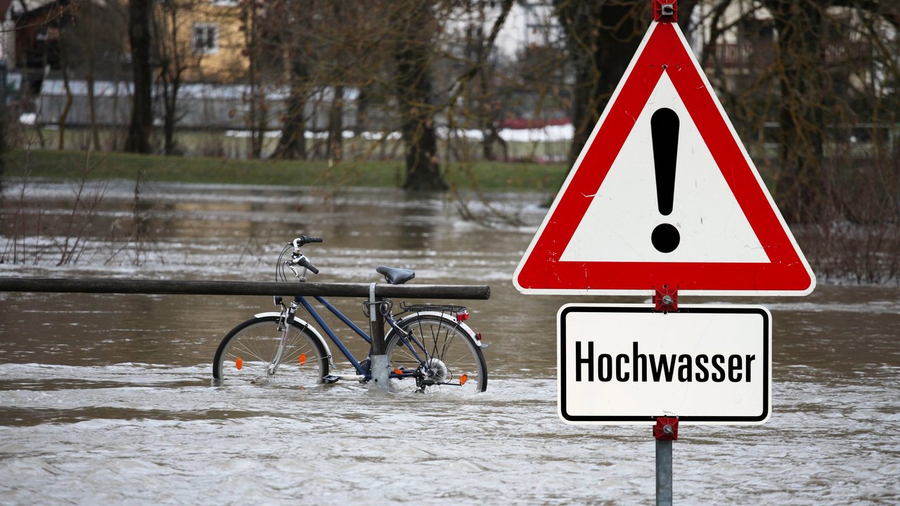 Gefahrenschild mit der Aufschrift "Hochwasser". Im Hintergrund sieht man Hochwasser mit einem Fahrrad