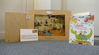 Ein Holzbuch steht aufgeklappt auf dem Boden, daneben ein Buch über die Olchis, davor ein kleines Buch