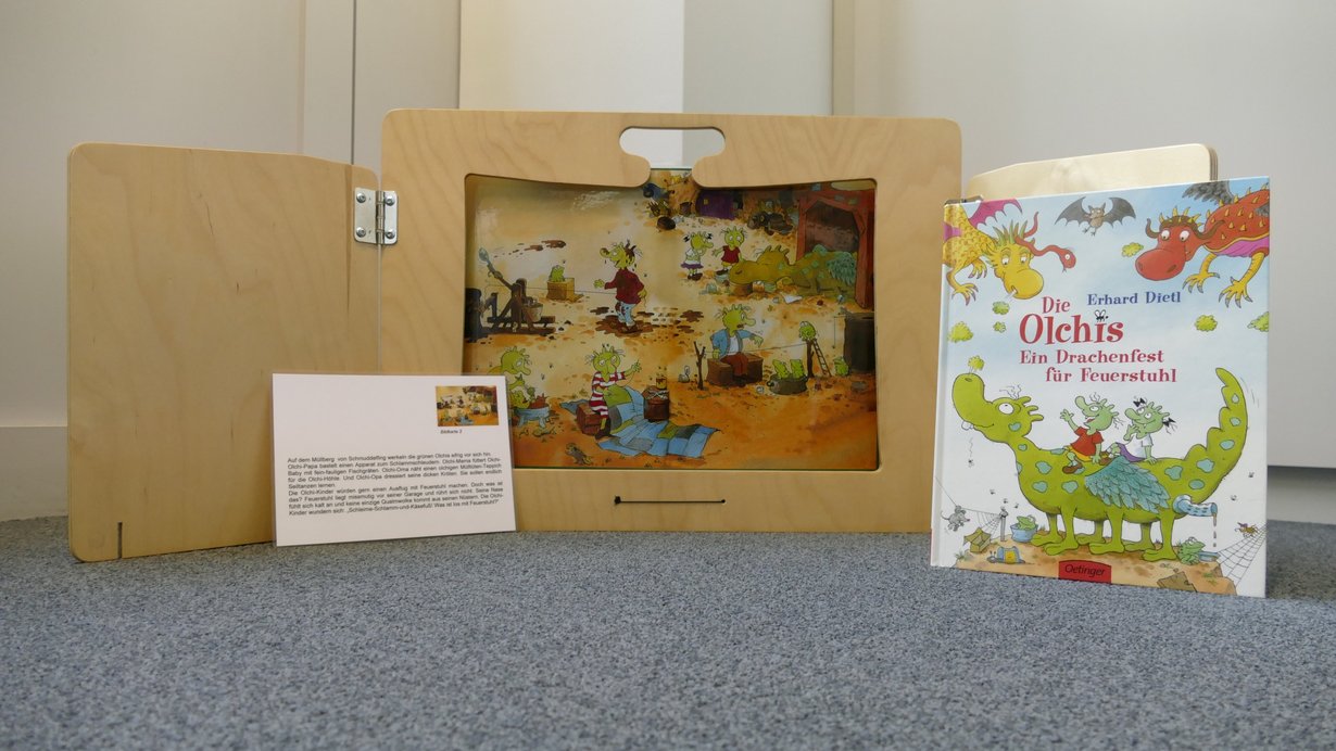 Ein Holzbuch steht aufgeklappt auf dem Boden, daneben ein Buch über die Olchis, davor ein kleines Buch