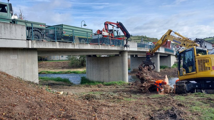 Arbeiter arbeiten mit einem Kran am Ufer eines Gewässer, auf der Brücke darüber stehen Baustellenfahrzeuge