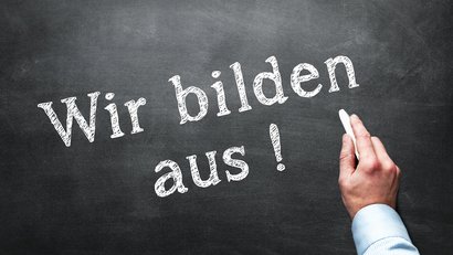 Bild zeigt Hand einer Person, die mit Kreide den Schriftzug "Wir bilden aus" an eine Tafel schreibt