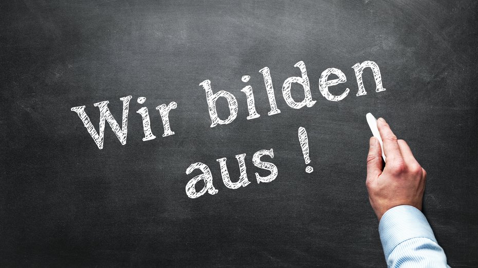 Schriftzug auf Tafel "Wir bilden aus" Bild zeigt Hand einer Person, die mit Kreide den Schriftzug "Wir bilden aus" an eine Tafel schreibt