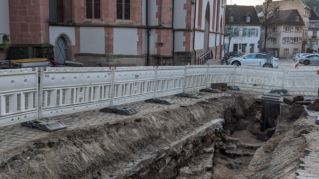 Blick auf die antiken Mauerzüge und Ziegel der römischen Unterbodenheizung, darüber moderne Verfüllungen und eine Betonplatte. Der Schnitt endet an einem modernen Thermalwasserschacht. Im Hintergrund der Marktplatz mit Stiftskirche.