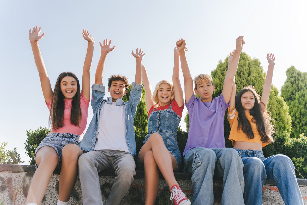 Mädchen und Jungen sitzen auf einer Mauer und strecken die Arme in die Höhe