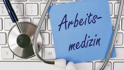 Bild zeigt Stethoskop mit Tastatur und einem Notizzettel mit der Aufschrift "Arbeitsmedizin"
