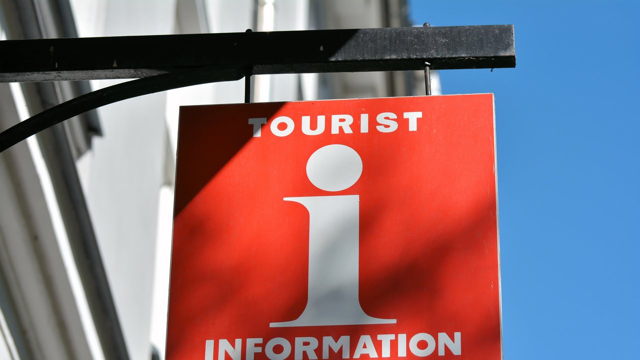 Rotes Schild an Hauswand mit weißem "i" in der Mitte oben "Tourist" unten "Information"