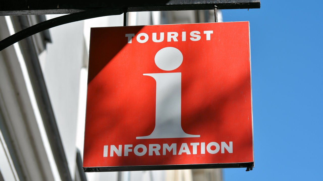 Rotes Schild an Hauswand mit weißem "i" in der Mitte oben "Tourist" unten "Information"