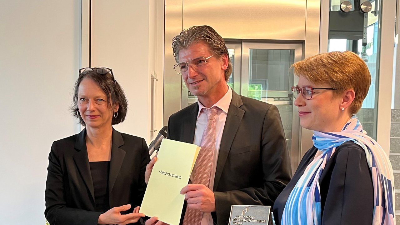 Bild zeigt Regierungspräsidentin Bay und Staatssekretärin Andrea Linklohr mit Bürgermeister Frank Menikheim. Dieser hält den Förderbescheid in den Händen