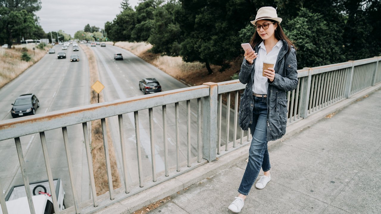 Eine Frau geht über eine Fußgängerbrücke, in einer Hand hält sie ein Handy, in der anderen einen Kaffeebecher