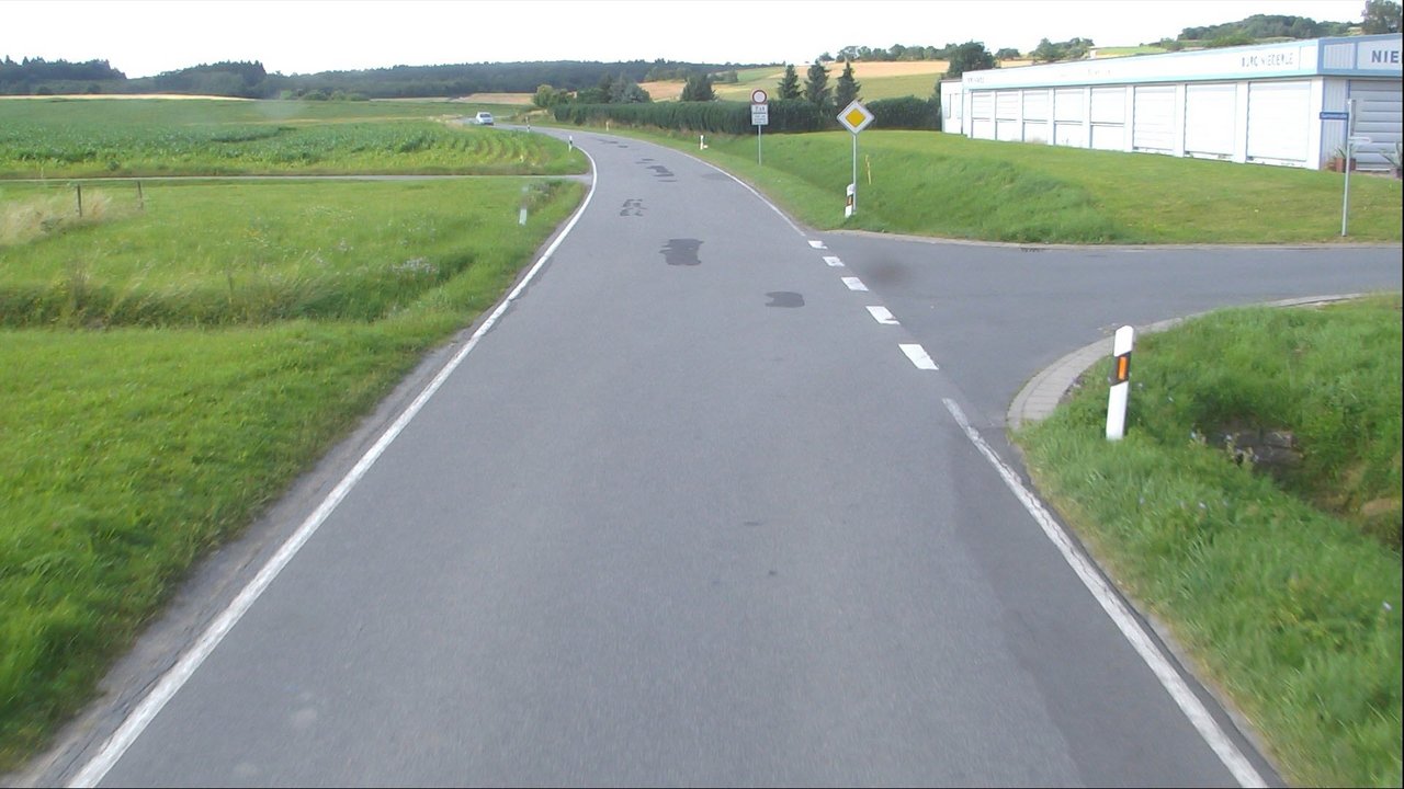 Bild zeigt eine Straße rechts und links davon Grünflächen, im Hintergrund ein Gebäude