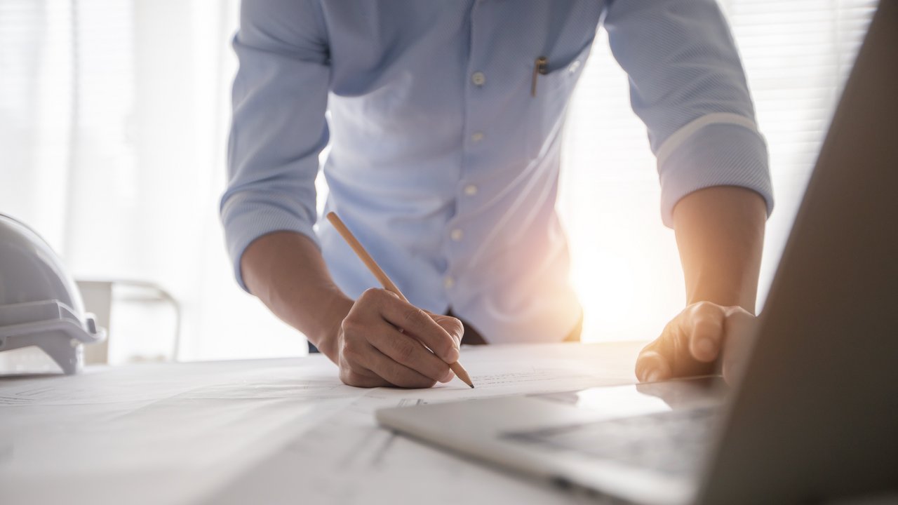 Ingenieur beugt sich über einen Plan und hat einen Stift in der Hand, im Vordergurnd sieht man einen Laptop