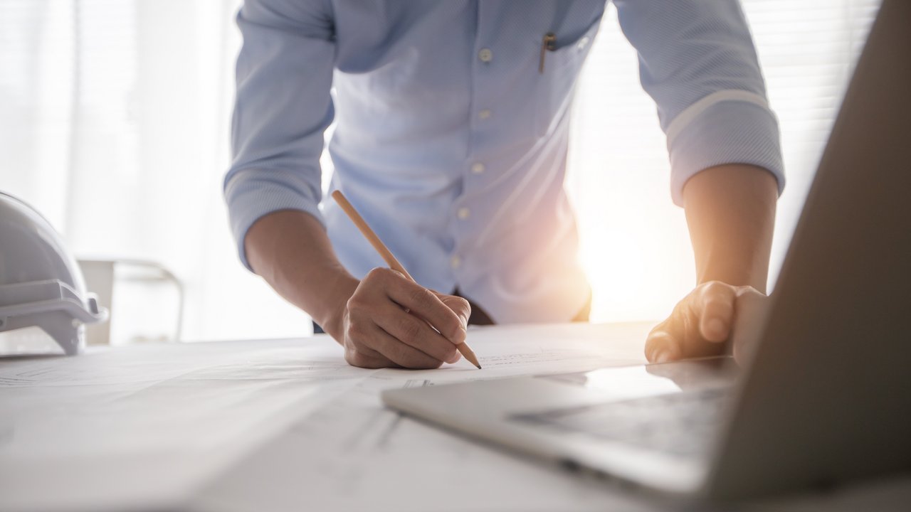 Ingenieur beugt sich über einen Plan und hat einen Stift in der Hand, im Vordergurnd sieht man einen Laptop