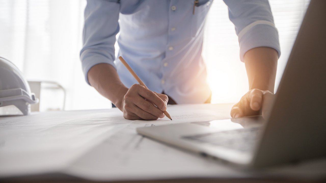 Ingenieur beugt sich über einen Plan und hat einen Stift in der Hand, im Vordergurnd sieht man einen Laptop